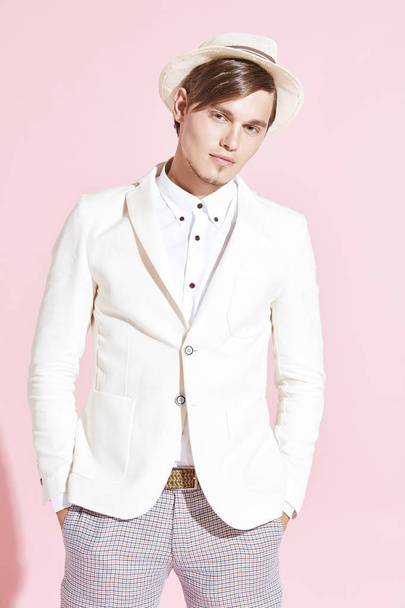 Portrait of young handsome serious modern man wearing white jacket, white shirt, grey pants and white hat posing in studio with light pink background - Zdjęcie, obraz