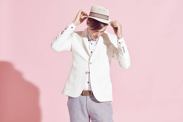 Portrait of young handsome serious modern male model wearing white jacket, white shirt, grey pants and white hat posing in studio with light pink background - Zdjęcie, obraz