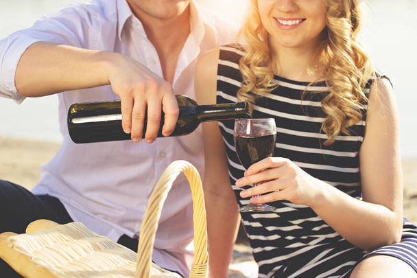 Enjoying picnic time together. Close-up of man in casual clothes pouring wine into glass while sitting on beach with his smiling girlfriend. - Φωτογραφία, εικόνα