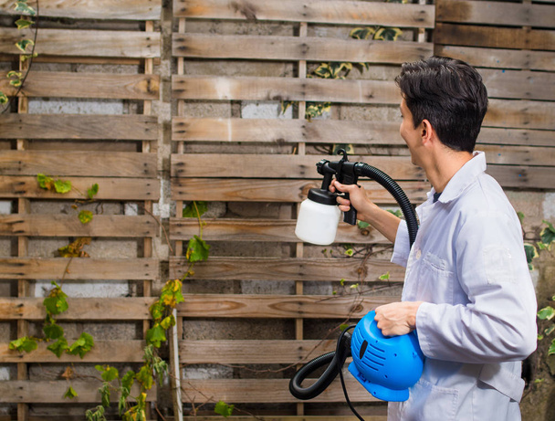 Trabalhador de construção bonito usando a pistola de pintura ao ar livre em uma estrutura de parede de madeira
 - Foto, Imagem