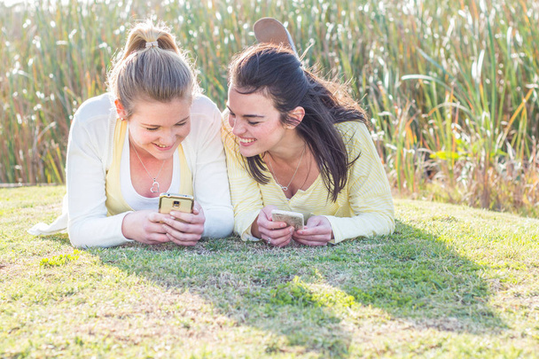 İki arkadaş elinde cep telefonu ile Park'ta rahatlatıcı çimenlerin üzerinde döşeme. - Fotoğraf, Görsel