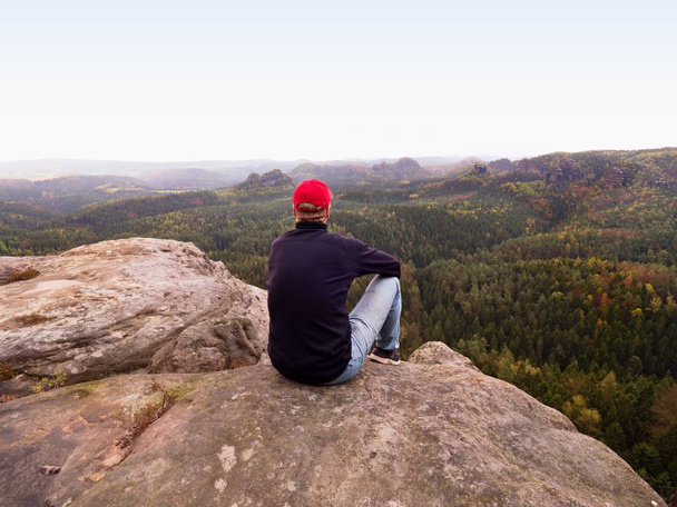 Turysta człowiek na klifie. Turystycznych w Czerwonym Kapturku, Czarna bluza i dżinsy siedzieć na ostre góry klif nad doliną - Zdjęcie, obraz