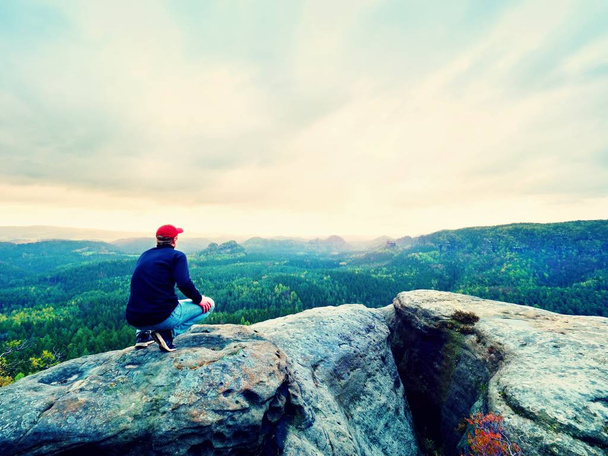 Tramp muž na skále. Turista v červené čepici, černý svetr a džíny sedí na ostré horské skále nad údolím - Fotografie, Obrázek