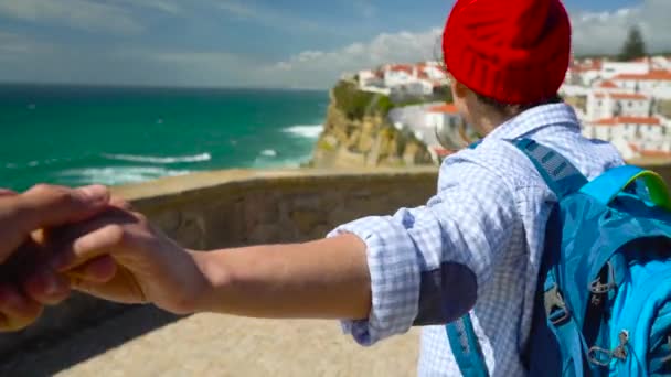 Sígueme - mujer joven feliz con un sombrero rojo y con una mochila detrás de su espalda tirando de chicos mano en Azenhas do Mar, Portugal. Caminando de la mano a la costa del océano
 - Imágenes, Vídeo