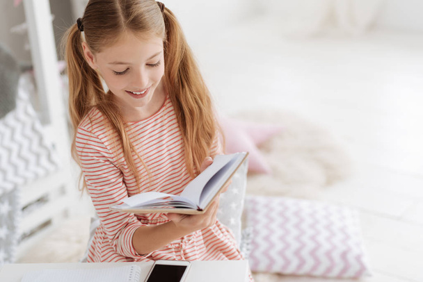 Smiling schoolgirl with ponytails reading book at home - Fotó, kép