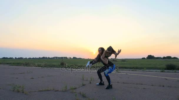 Beautiful, athletic, young woman in sunglasses, in tights, performs squats, holding a girl in her arms, exercises in pairs, as an additional weight. on an abandoned airfield, sunset - Footage, Video