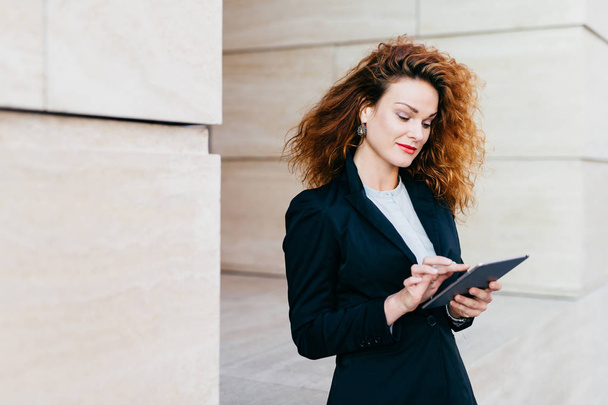 Portrait of young attractive female entrepreneur working on new business project using electronic gadget, having pleased look into screen. Curly woman in black costume standing outdoor near builduing - Photo, Image