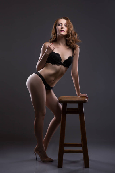 Sexy and beautiful young woman withwavy hair, long legs and bronzed skin is posing in the black lacy underwear in the studio on the chair, dark background - Zdjęcie, obraz