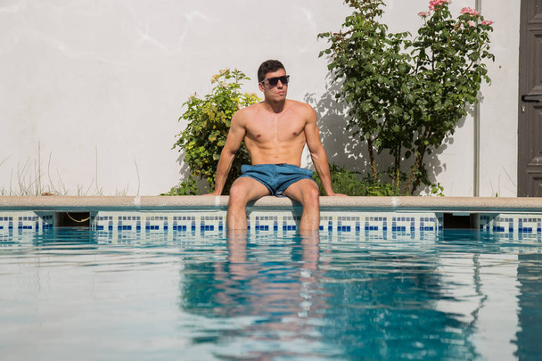 Vahva nuori mies istuu uima-altaan reunalla aurinkoisena päivänä
  - Valokuva, kuva