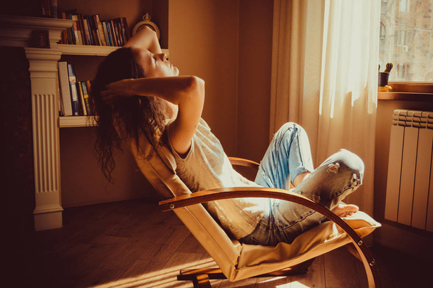 Woman relaxing after work in comfortable modern chair near window in livingroom. Warm natural light. Cozy home. Casual clothing. Casual style indoor. Room interior. Tired and thoughtful woman. - Zdjęcie, obraz