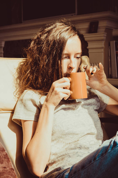 家庭での時間を楽しんでいます。クローズ アップ肖像画栗髪の女性がお茶やコーヒーを飲むウィンドウで快適な椅子でリラックス。自然光。居心地の良い家。瞬間をお楽しみください。明るく晴れた日。カップのホット飲料 - 写真・画像