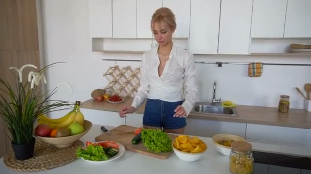 Slanke blonde vrouw voltooit voorbereiding van gezond ontbijt en verspreidt groenten, permanent aan tafel van moderne keuken. - Video