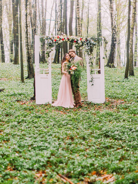 The lovely photo of the newlywed couple in the vintage clothes standing head-to-head to each other under the white arch in the form of two doors decorated with colourful flowers in the green wood. - Zdjęcie, obraz
