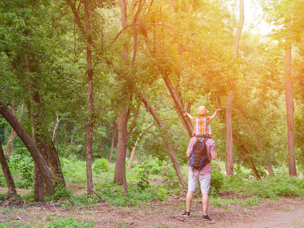 Νεαρός πατέρας με τον γιο στους ώμους στέκεται με φόντο πράσινο δάσος. Πίσω όψη. Ηλιακό φως. - Φωτογραφία, εικόνα