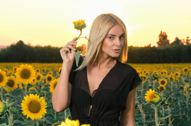 Portret prachtige jonge blonde model in zwarte jurk op een veld met zonnebloemen - Foto, afbeelding