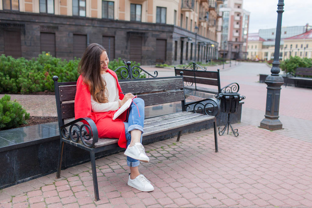 Uma garota atraente com cabelos castanhos longos e um sorriso de dente branco se senta em um banco e escreve seus pensamentos sobre o fundo urbano em um caderno vermelho. Ela está vestindo uma camisola branca e jeans azul
 - Foto, Imagem