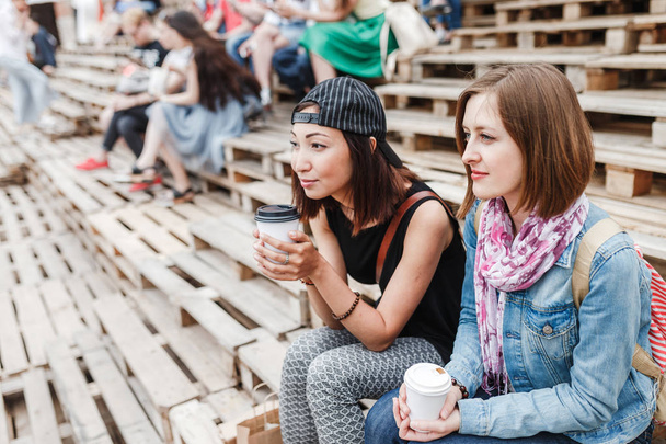 Dvě roztomilé mladé ženy s kávou s sebou sedí na lavičce, mluví a sledování venkovní pouliční představení - Fotografie, Obrázek