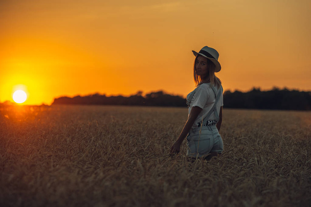 Молодая девушка с распростертыми руками стоит на пшеничном поле на закате - Фото, изображение