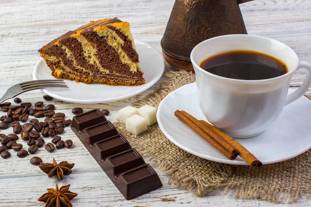 Delicious chocolate cake on a plate with a cup of coffee . Coffee beans, a piece of chocolate, anice, sugar and cinnamon sticks on wooden table - Photo, Image