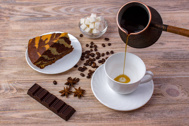 Κάτοψη του ένα φλιτζάνι καφέ και ένα κομμάτι της νόστιμο κέικ σε ένα πιατάκι, κόκκους καφέ, ένα μπολ με ζάχαρη σε κύβους, σοκολάτα και γλυκάνισο στο φωτεινό ξύλινο τραπέζι - Φωτογραφία, εικόνα
