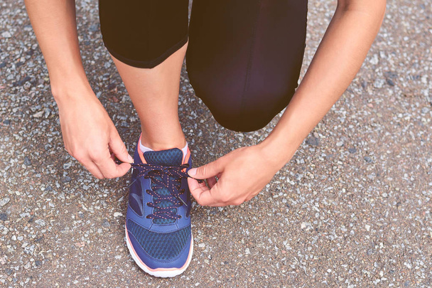 Girl ties up shoelaces in sneakers on road while jogging, legs and sneakers, toned image - Fotó, kép