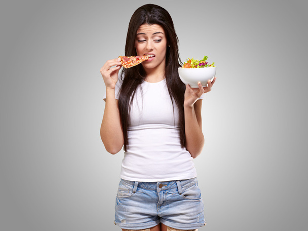 portrait of young woman eating pizza and looking salad over grey - Photo, Image