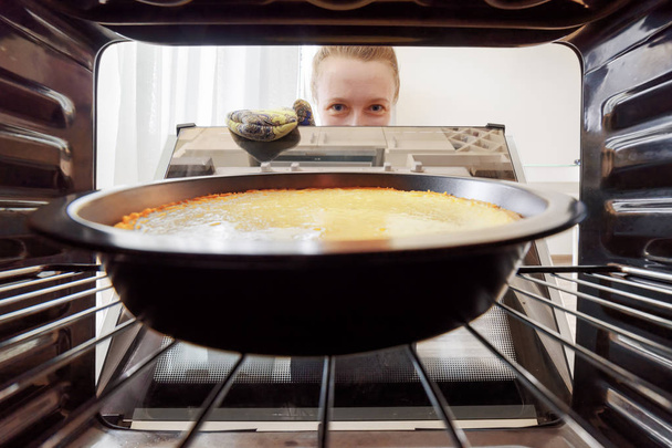 Young housewife looking at cheesecake into oven in kitchen - 写真・画像
