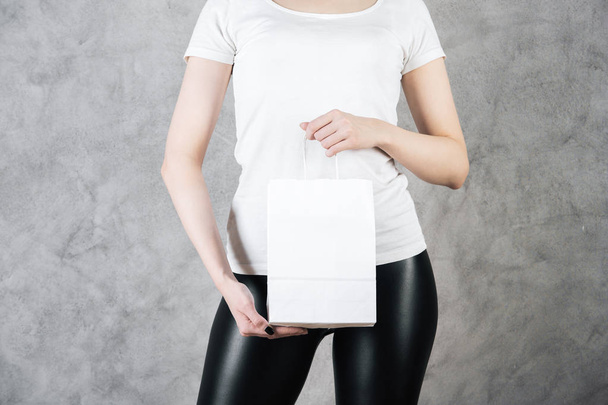 Close up of woman holding empty white paper bag for shopping on concrete wall background. Supermarket concept. Mock up  - Fotoğraf, Görsel