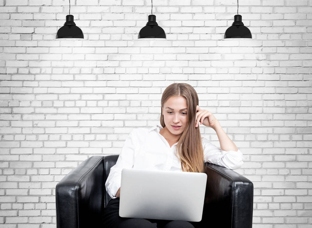 Blonde woman in armchair, laptop, brick - Zdjęcie, obraz