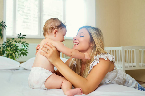 Mother and baby in a diaper play hugging on a bed indoors.  - Photo, image