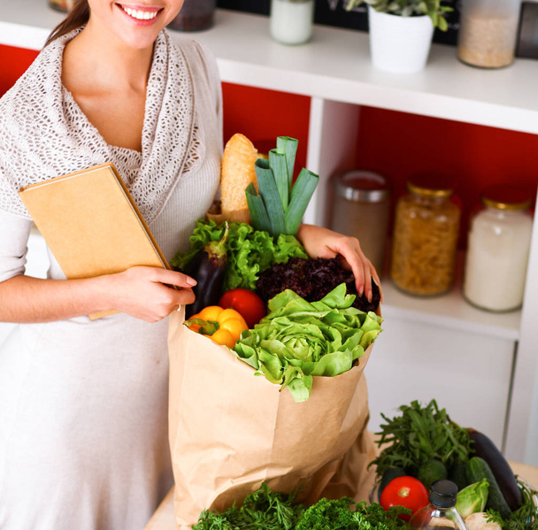 Młoda kobieta trzyma torbę na zakupy z warzywami Stojąc w kuchni - Zdjęcie, obraz