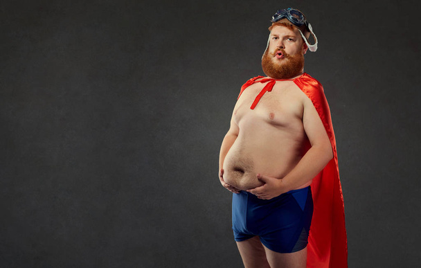Fat funny man in swimming trunks in a superhero costume holds on - Фото, зображення