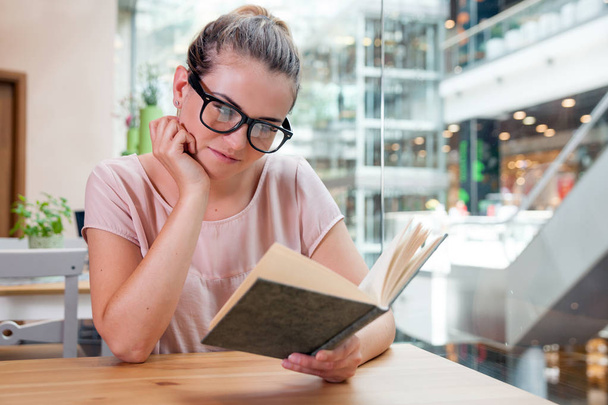Relajada chica sonriente leyendo libro en la cafetería en el centro comercial
 - Foto, Imagen