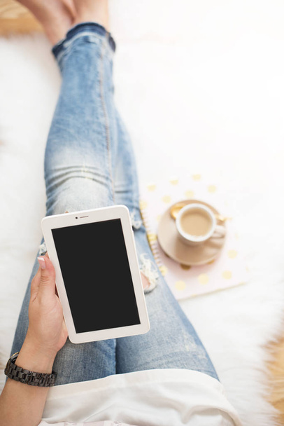 A young woman wearing distressed jeans sitting on wood floor on a white fur carpet at home and holds a tablet in his hand. Around a cup of coffee, notebooks. Gold bright feminine theme - Φωτογραφία, εικόνα