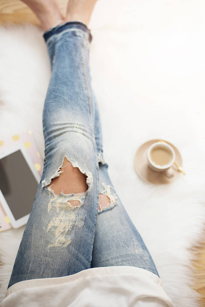 A young woman wearing distressed jeans sitting on wood floor on a white fur carpet at home. Around a cup of coffee, tablet and notebooks. Gold bright feminine theme - Foto, immagini