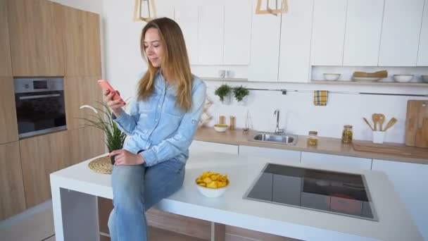 Beautiful woman uses cell phone for correspondence and tries potato chips sitting on surface of kitchen table. - Filmati, video