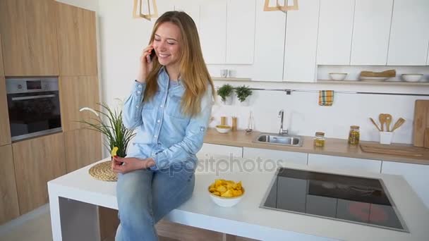 Krásná mladá žena chatování na telefonu a brambůrek sedí na povrchu kuchyňského stolu. - Záběry, video