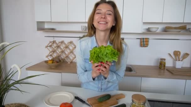 Gelukkig vrouwelijke huisvrouw neemt stelletje salade verlaat in handen en vormt met glimlach op camera, staande in het midden van de keuken op de keukentafel in ochtend. - Video