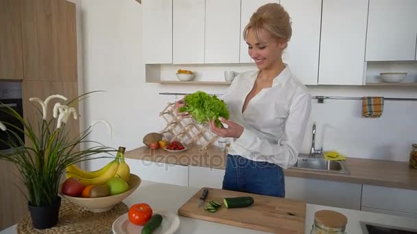 Όμορφη νοικοκυρά γυναίκα κρατώντας φύλλα πράσινη μαρούλι και χαμογελώντας, ενώ στέκεται στο τραπέζι της κουζίνας, μοντέρνα κουζίνα. - Πλάνα, βίντεο