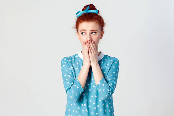 The confused young girl. girl covering mouth with both hands keeping a secret. Beautiful redhead girl in blue dress doesn't want to spread rumors or some confidential information. Isolated studio shot on gray background. - Photo, Image