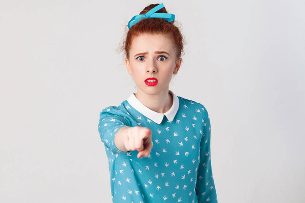The seriosly redhead girl, wearing blue dress, opening mouths widely, having surprised shocked looks, pointing finger at camera. Isolated studio shot on gray background - Photo, Image