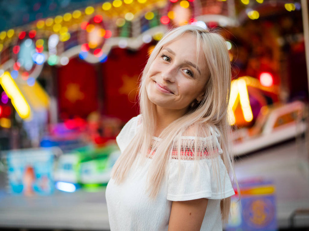 Young attractive blonde girl standing near carousel in amusement park. Beautiful portrait of woman with long hair. - Foto, Bild