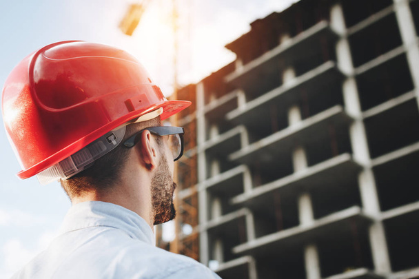 Young engineer in red helmet inspects construction of building. Bearded businessman on background of skyscraper under construction. Portrait of construction engineer on construction site background - Photo, Image