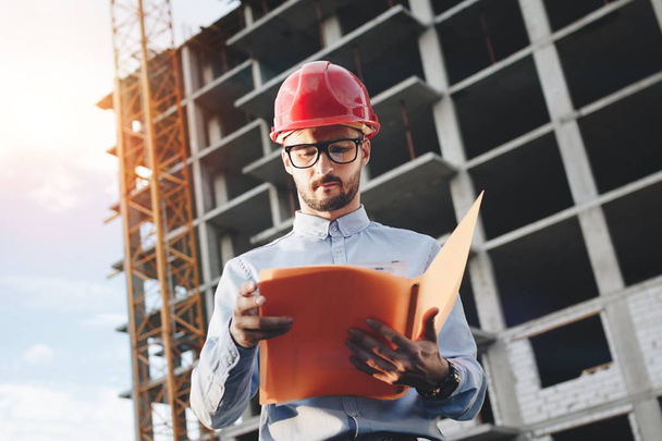 Young bearded engineer in red construction helmet on background of a building under construction with drawings and a folder in his hands - Valokuva, kuva