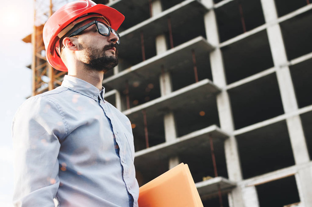 Young bearded businessman or engineer wearing glasses and helmet on construction site. Portrait of construction inspector with folder in hand on building construction background - Foto, immagini
