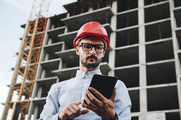 Young business man builder in red hard hat with tablet in hand. Portrait engineer or architect with tablet on construction site - Foto, immagini