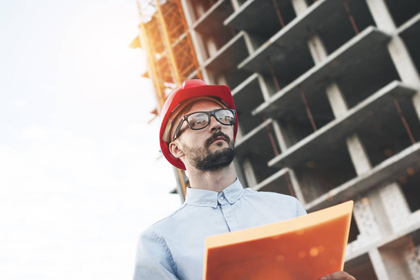 Portrait of modern male engineer at construction site of new modern plant. Inspector holds a folder with documents and checks the progress of the building construction. Lens flare effetc, blurred background - Фото, изображение