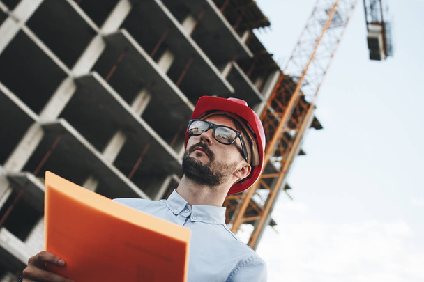 Young modern engineer builder or architect with folder of documents on background of building being built and crane. Industrial Safety Inspector checks progress of construction of industrial enterprise - Fotó, kép