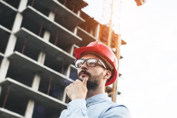 Thoughtful engineer in red hard hat thinks over construction building. Portrait of architect in glasses solving problems at construction site - Foto, imagen