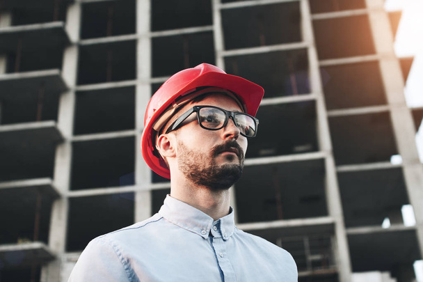 Young successful businessman builder with glasses and red hard hat on construction site inspects construction of new modern factory - Фото, изображение